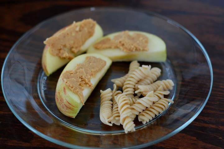 Three apple slices with peanut butter on them and a side of spiral noodles on a plate for a snack.