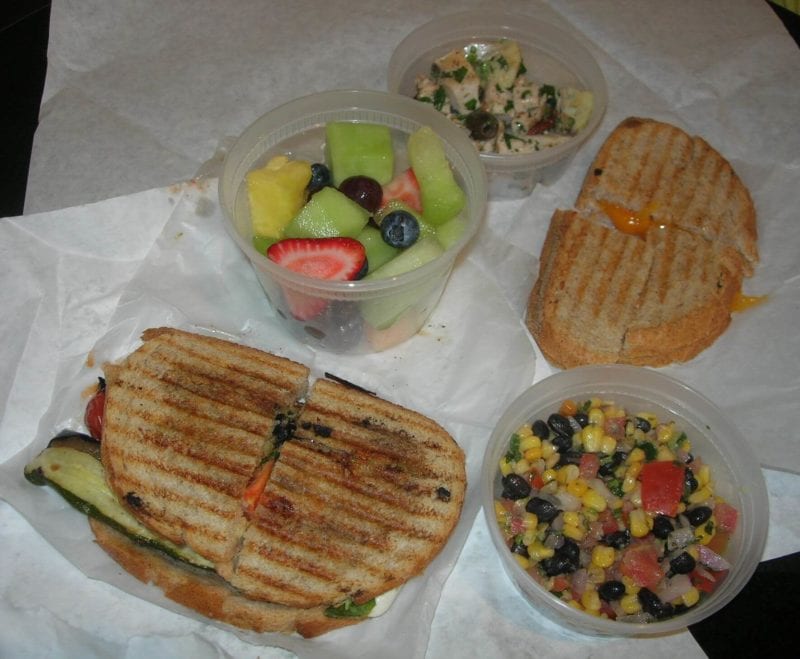 A variety of food ordered from a restaurant that includes a Mediterranean grilled vegetable and mozzarella sandwich, fresh fruit, grilled cheese sandwich, Greek chicken salad, and corn/black bean/tomato salad. 