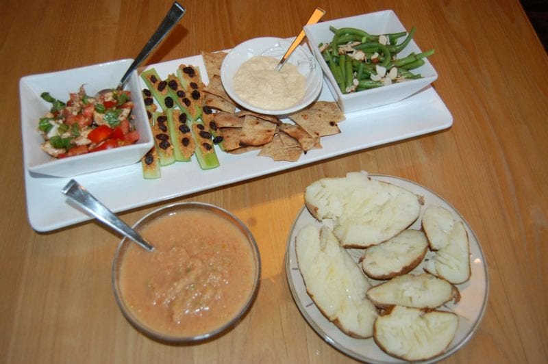 Crackers and hummus, baked potatoes, green beans, and a bowl of gazpacho plated on the dinner table. 