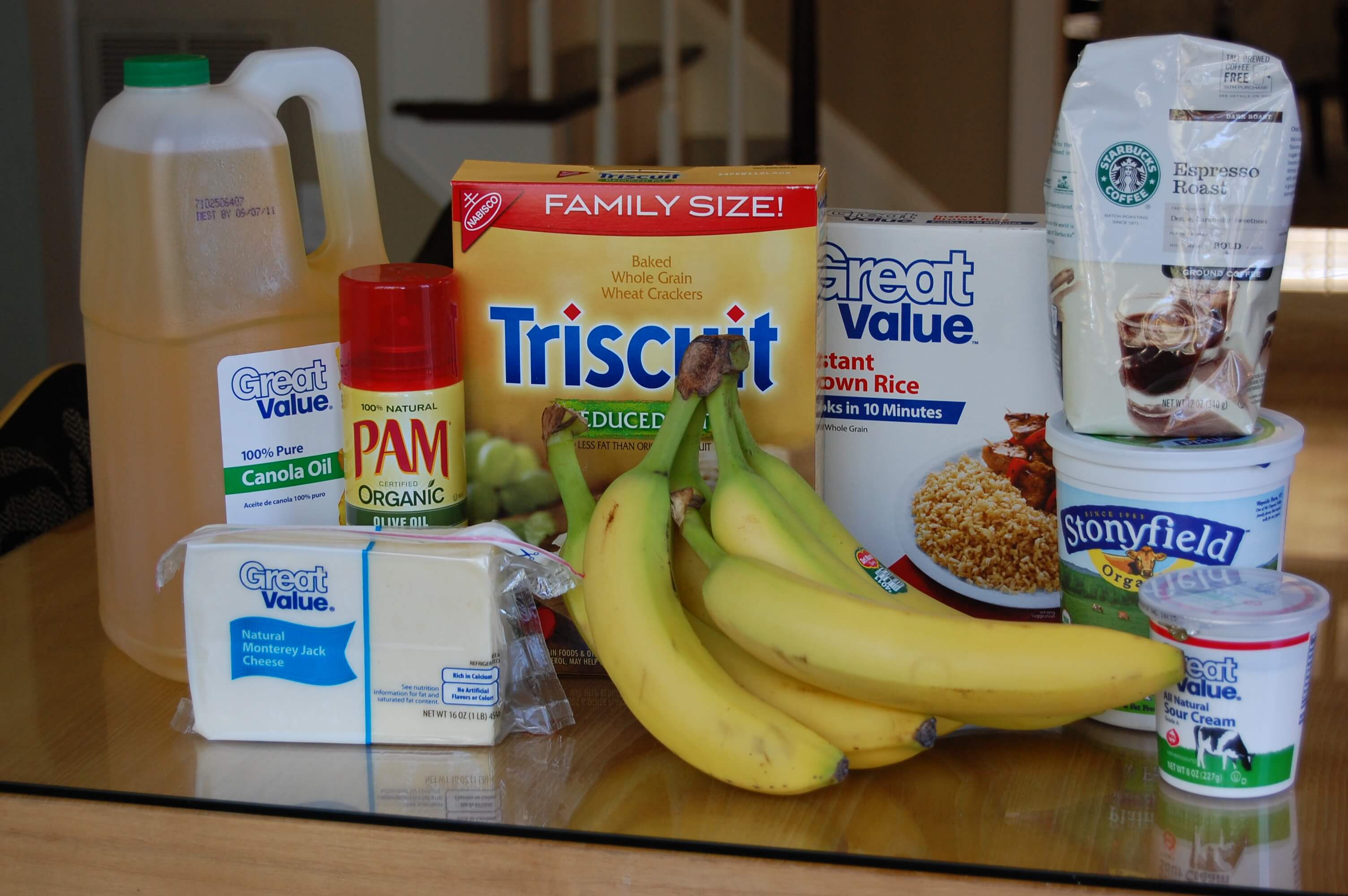 Groceries from Wal-Mart on a table that include coffee, bananas, cheese, Triscuits, brown rice, yogurt, sour cream, and cooking oil. 