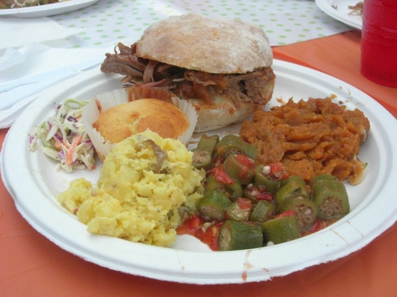 Plate of pulled pork on a bun, potato salad, corn bread muffin, coleslaw, and mashed sweet potatoes. 