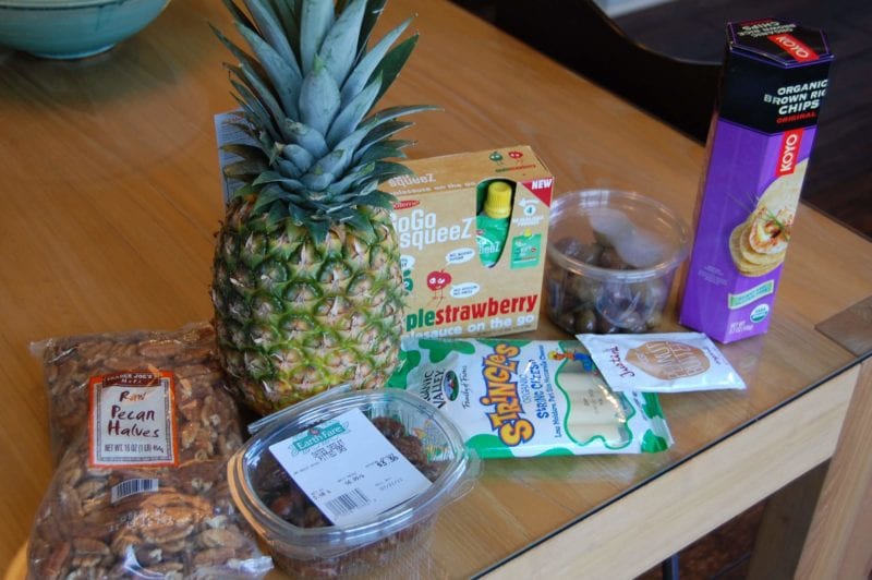Groceries from Earth Fare that include pineapple, string cheese, pecans, dates, Squeeze applesauce, peanut butter, olives, and brown rice crackers. 