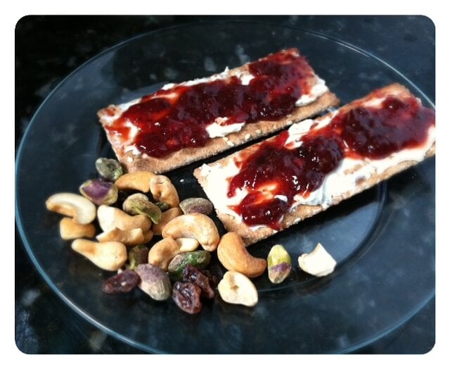 Whole grain snack crackers topped with strawberry jam and cream cheese, and a handful of mixed nuts on a plate.